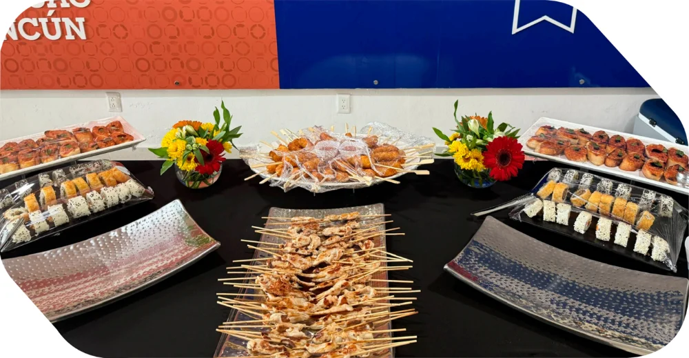 Barra de bocadillos y canapés con rollos de sushi, brochetas y centros de mesa florales para eventos en Cancún.